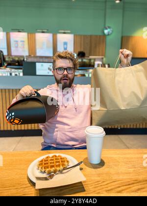 Ein überraschter Mann sitzt mit einer Waffel an einem Café-Tisch, hält eine große Einkaufstasche und eine Geschenkbox zum Mitnehmen. Moment der unerwarteten Freude und des Genusses in einem Stockfoto