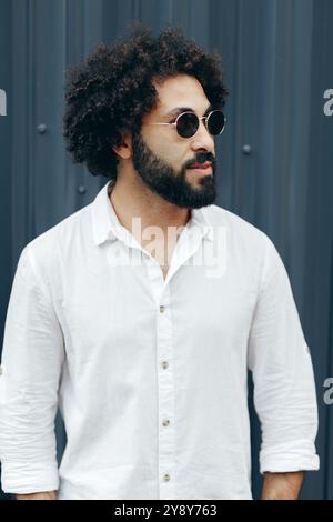 Ein arabischer Mann mit Afro-Frisur und Bart in weißem Hemd, der an der Metallprofilwand steht Stockfoto
