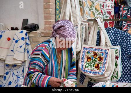 Buchara, Usbekistan; 19. September 2024: Eine ältere usbekische Frau verkauft geschickt exquisite Seidenstücke an faszinierte Touristen in der pulsierenden Stadt Buk Stockfoto