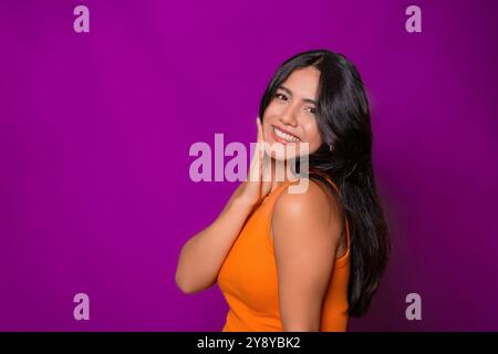Fröhliche Frau in lässigem Orange Oberteil, Hand auf Gesicht, lächelnd warm, lila Hintergrund Stockfoto