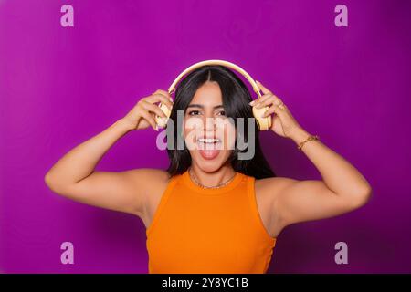 Frau in einem orangefarbenen Oberteil mit gelben Kopfhörern, lächelt und hält die Kopfhörer auf beiden Ohren und zeigt Aufregung vor einem leuchtend violetten Hintergrund. Stockfoto
