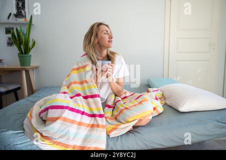 Eine entspannte Frau mittleren Alters, die in eine Decke gehüllt ist und eine Tasse Morgenkaffee im Bett hält, blickt ins Fenster. Stockfoto