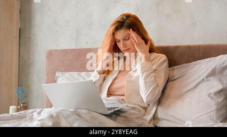 Freiberuflicher Kaukasische Frau arbeitet fernab von zu Hause im Bett denkend nachdenklich, Kopf mit Laptop-Arbeit im Schlafzimmer schlecht Kopfschmerzen zu fühlen Stockfoto