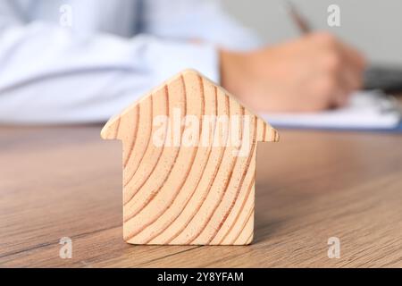 Immobilienversicherung. Ein Mann schreibt etwas am Tisch, konzentriert sich auf eine hölzerne Hausfigur Stockfoto