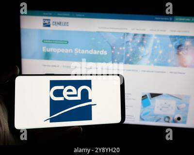 Person, die ein Mobiltelefon mit dem Logo des Europäischen Komitees für Normung (CEN) auf dem Bildschirm vor der Webseite hält. Schwerpunkt auf dem Display des Telefons. Stockfoto
