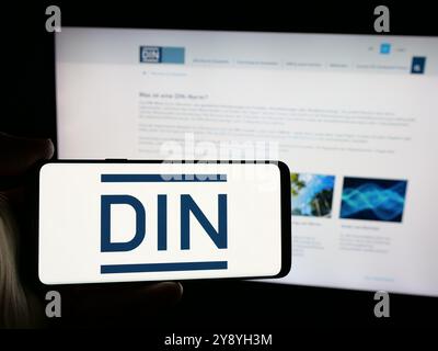 Person mit Smartphone und Logo des Deutschen Instituts für Normung e.V. (DIN) auf dem Bildschirm vor der Website. Schwerpunkt auf dem Display des Telefons. Stockfoto