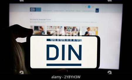 Person mit Handy mit Logo des Deutschen Instituts für Normung e.V. (DIN) auf dem Bildschirm vor der Webseite. Schwerpunkt auf dem Display des Telefons. Stockfoto