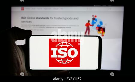 Person mit Mobiltelefon und Logo der Internationalen Organisation für Normung (ISO) auf dem Bildschirm vor der Webseite. Schwerpunkt auf dem Display des Telefons. Stockfoto