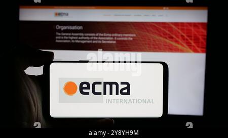 Person mit Mobiltelefon und Logo der Normungsorganisation Ecma International auf dem Bildschirm vor der Webseite. Schwerpunkt auf dem Display des Telefons. Stockfoto