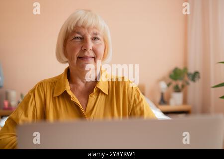 Glückliche Seniorin, die bei der Arbeit zu Hause mit einem Laptop in die Kamera schaut. Schöne, stilvolle ältere Dame, die ihre Website auf dem Handy durchsucht. Stockfoto