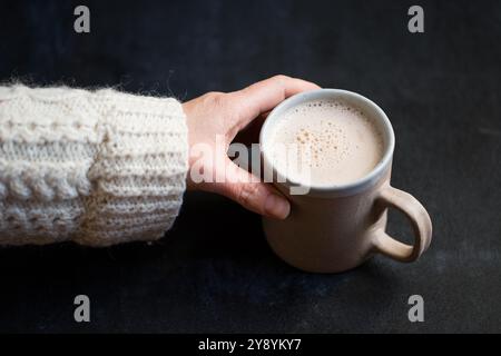 Frau, die eine Tasse heiße Schokolade oder Kaffee hält, kuscheliges Wintergetränk in Keramikbecher, warmer Strickpullover und Lebensstil im dunklen Hintergrund. Stockfoto