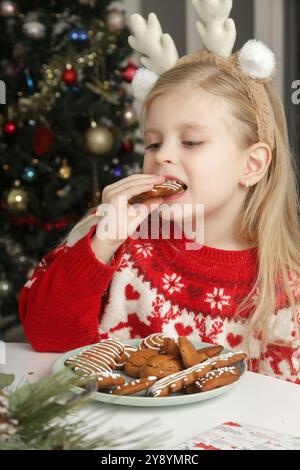 Entzückendes kleines Mädchen, das zu Hause Winterferien genießt, Weihnachtspullover trägt, Lebkuchenkekse auf dem Tisch, gemütliche festliche Atmosphäre. Stockfoto