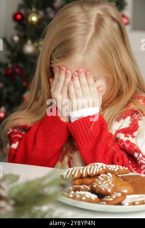 Trauriges, frustriertes kleines Mädchen im Weihnachtsoutfit und Weihnachtsmann-Hut am Tisch mit Lebkuchenkeksen, Winterurlaub-Stress-Konzept. Stockfoto