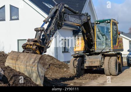 Ein schweres Baufahrzeug gräbt aktiv in der Nähe eines zeitgenössischen Hauses, was darauf hinweist, dass in einem Wohngebiet unter klarem Himmel laufende Entwicklungsarbeiten durchgeführt werden Stockfoto