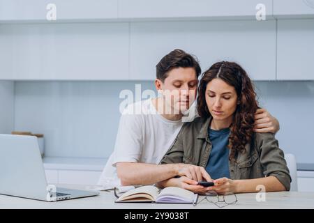 Verheiratetes Paar, das zu Hause gemeinsame Budgetierung macht, mit Rechner, Smartphone und Laptop, Mann unterstützt Frau, indem er ihre Schulter umarmt. Familienförderung in der Finanzkompetenz Stockfoto