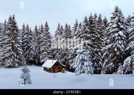 Märchenweihnachtslandschaft mit leuchtendem Holzhaus in verschneiten Wäldern. Gemütliches Haus in den Winterbergen Stockfoto