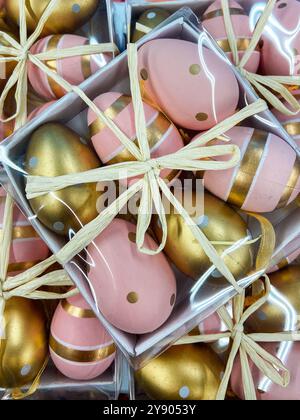 Auswahl an dekorativen Pastelleiern und goldenen Eiern für Ostern, ordentlich organisiert in transparenten Kästen, gebunden mit rustikalen Schnur. Die festliche Anordnung erinnert an eine Stockfoto