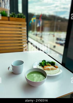 Gesunde Mahlzeit mit einer Schüssel grüner Suppe und einem Teller mit Brokkoli und Fisch wird auf einem weißen Tisch durch ein großes Fenster gestellt. Friedliche Atmosphäre Stockfoto