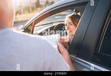 Junge Frau sitzt auf dem Autositz und lächelt aufrichtig zum Liefermann und nimmt zwei Pizzaschachteln auf der Straße der Stadt an. Kleine Unternehmen, Menschen Rela Stockfoto