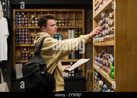 Seitenansicht eines jungen Mannes in beigefarbenem Hoodie, der vor einem großen Display mit Graffiti-Farben steht, während er eine Dose mit definierter Farbe wählt Stockfoto