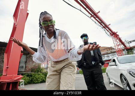 Frau mit Zöpfen und grüner Sonnenbrille, die eine Geste mit den Händen macht, Mann in schwarzem Outfit, der unter industrieller Struktur im Hintergrund steht Stockfoto