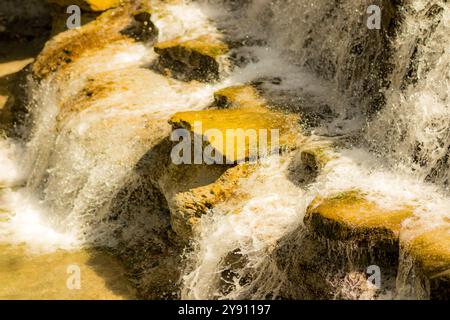 Ein kleiner Wasserfall, der über zerklüftete Felsen kaskadiert. Das Bild gleicht die warmen Erdtöne der Felsoberfläche mit dem kühlen, schaumigen Rauschen des Wassers aus. Stockfoto