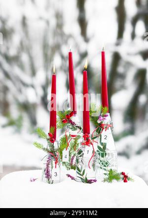 Adventskranz mit vier rot brennenden Kerzen und weißen hölzernen Zahlenanhängern in Glasflaschen, die mit Tannenzweigen verziert sind. Stockfoto