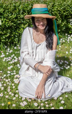Frau mit Sonnenhut, die auf einem Feld von Gänseblümchen sitzt und die Schönheit des Sommers im Freien genießt, Glückskonzept Stockfoto
