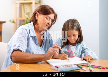 Fokussierte Oma und Enkelin malen im Buch zusammen auf dem Tisch zu Hause. Die Szene ist von Kreativität und Wärme erfüllt, während sie sich über ihre künstlerische Tätigkeit vereinen Stockfoto