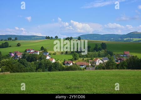 Kleines Dorf eingebettet in eine hügelige Landschaft mit grünen Wiesen und Bäumen unter blauem Himmel, Wachtkueppel, Gersfeld, Rhoen, Hessen, Deutschland, Europa Stockfoto