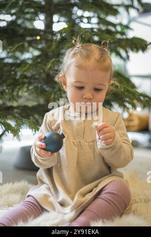 Kleinkind in einem beigefarbenen Kleid, das eine Weihnachtsbaumkugel vor einem festlich geschmückten Baum hält Stockfoto