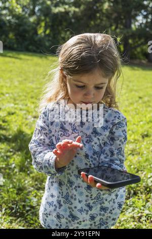 Ein kleines Mädchen hält ein Smartphone und steht nachdenklich auf einer sonnigen Wiese Stockfoto