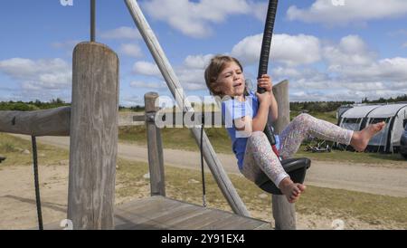 Ein Kind springt fröhlich auf eine Seilrutsche in einem weiten, sonnigen Spielplatz mit bewölktem, blauem Himmel Stockfoto