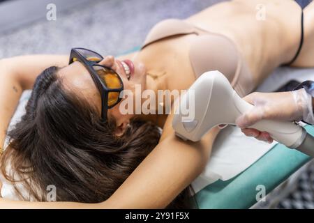 Frau in Protect Brille, die auf einer Couch liegt und eine Laser-Haarentfernung für Achselhöhlen bekommt Stockfoto