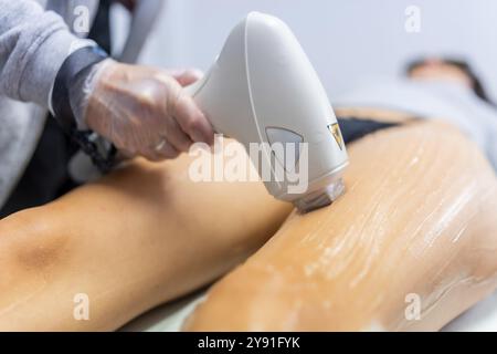 Nahaufnahme einer kaukasischen Ärztin, die ein Kosmetologiegerät hält, während die Frau, die auf dem Bett liegt, mit Laser-Machin eine Haarentfernung der Wadenbeine erhält Stockfoto