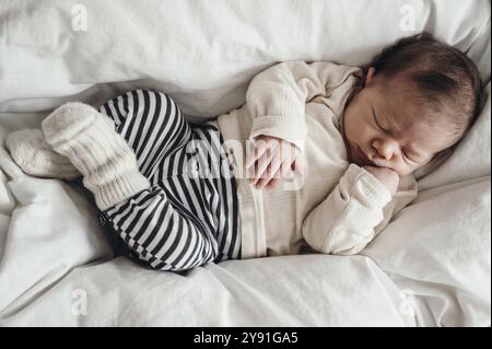 Schlafendes Baby in einem gestreiften Strampler, das auf einem weißen Bett liegt, sieht friedlich und sicher aus Stockfoto