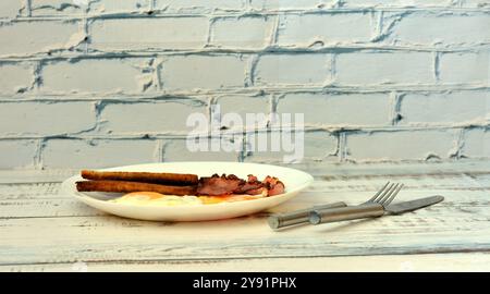 Ein Teller mit zwei Spiegeleiern, knusprigen Speckscheiben und Toast auf einem hellen Holztisch, Besteck in der Nähe. Nahaufnahme. Stockfoto