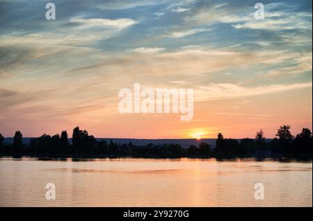 Der atemberaubende Sonnenuntergang erleuchtet den Himmel in Rosa, Orange und Blau und strahlt warmes Licht über dem ruhigen See aus. Silhouettenbäume und weit entfernte Häuser säumen das Ufer und schaffen eine malerische und ruhige Szene. Stockfoto