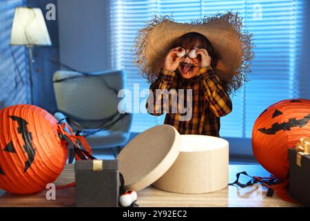 Süßer Junge, der wie Vogelscheuche gekleidet ist, mit dekorativen Augäpfeln, festlicher Dekoration und Geschenkboxen drinnen in der Nacht. Halloween-Feier Stockfoto