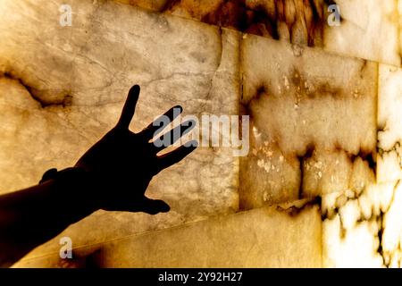 Eine Hand greift nach außen, um eine Wand mit einer Steinoberfläche zu berühren. Die Hand ist im Schatten, und die Wand ist mit einem Muster aus Stein bedeckt. Die Szene ist eine von Cur Stockfoto