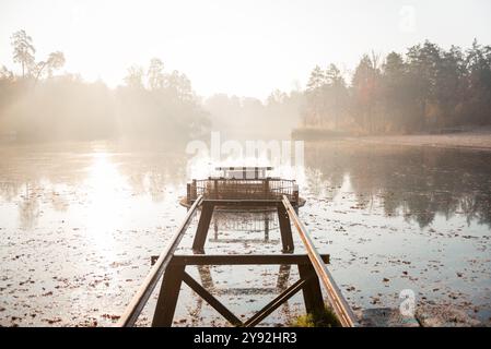 Morgens auf dem See. Metallschienen, die zu einem runden Überlauf führen. Wald bedeckt mit Nebel im Hintergrund, ruhiges Wasser reflektiert die Bäume. Stockfoto