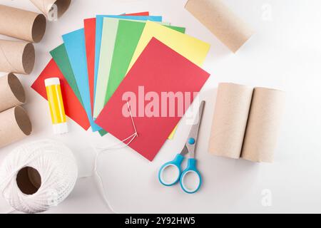 Bastelmaterialien für Papierdekorationen, mit farbigen Papierbögen, Pappröhrchen, Klebestift, Schere, Nadel, und weißes Gewinde. Flache Lay-Komposition für Heimwerker und Bastelprojekte. Stockfoto