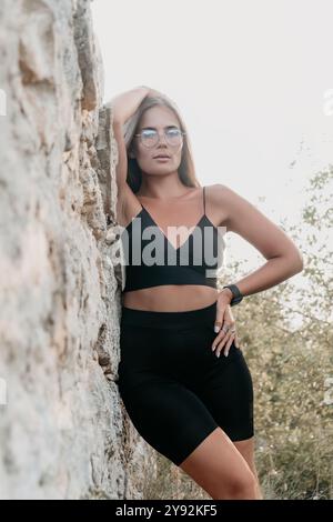 Frau, Felsen, draußen: Eine Frau, die eine Brille und schwarze Sportkleidung trägt, während sie sich in einer natürlichen Umgebung an eine Felsformation lehnt. Stockfoto