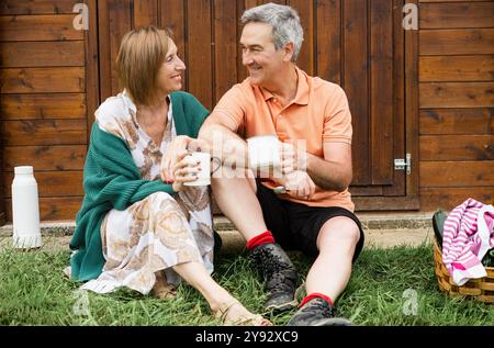 Lächelndes Paar, das auf Gras sitzt und Kaffee in ländlicher Umgebung genießt. Stockfoto