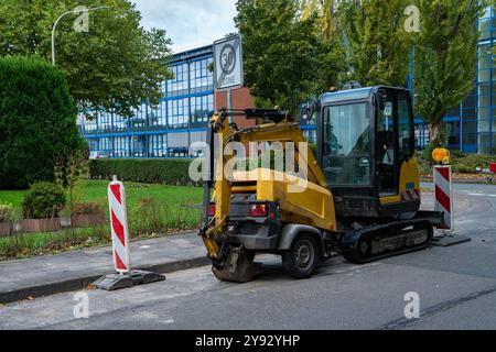 Ein kleiner gelber Baubagger parkt auf einer Stadtstraße in der Nähe eines Universitätsgeländes. Verkehrsschilder und Barrieren weisen auf laufende Straßenarbeiten in der Gegend am Nachmittag hin. Stockfoto