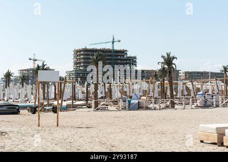 Leerer Strand mit zusammengefalteten Liegestühlen und Sonnenschirmen, vor dem Hintergrund einer Baustelle mit Kränen und einem teilweise erbauten mehrstöckigen Gebäude Stockfoto