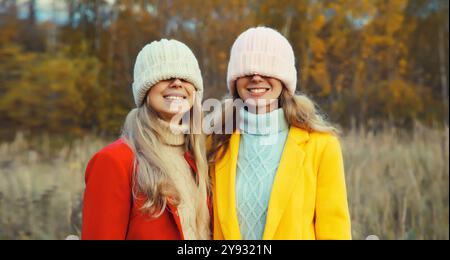 Glückliche zwei Freundinnen ziehen gestrickte Wintermütze über ihr Gesicht, die ihre Augen bedeckt, lustige Freundinnen, die sich im Park amüsieren Stockfoto