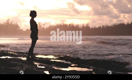 Frau schlanke Silhouette stehend felsiger Strand, Ozean dehnt sich aus. Wasserwellen krachen unter Sonnenuntergang gegen die Felsen. Stockfoto