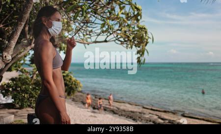 Eine Frau mit Gesichtsmaske steht selbstbewusst am Sandstrand, die Wellen streicheln sanft das Ufer hinter sich. Trotz der Pandemie findet sie Trost und Frieden in der Schönheit des Ozeans. Stockfoto