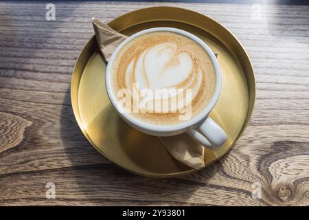 Cappuccino mit Herz Latte Art auf Holztisch in goldener Untertasse im Café. Stockfoto